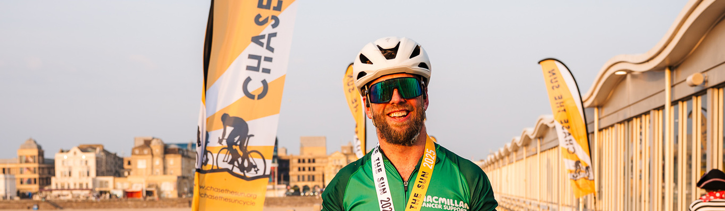 A cyclist is standing on a beach pier propping up a bicycle with their right arm. They are wearing cycling gear including a green Macmillan branded cycling top. They also have a Chase the Sun finishers medal around their neck. 