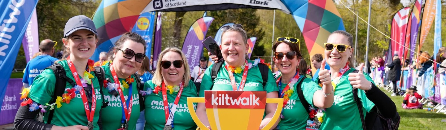 A group of event participants are standing in a row. They are all wearing green Macmillan branded tops and red kilts. One person is holding up a paper cut out of a trophy. They are standing in front of a large event finish balloon arch. 
