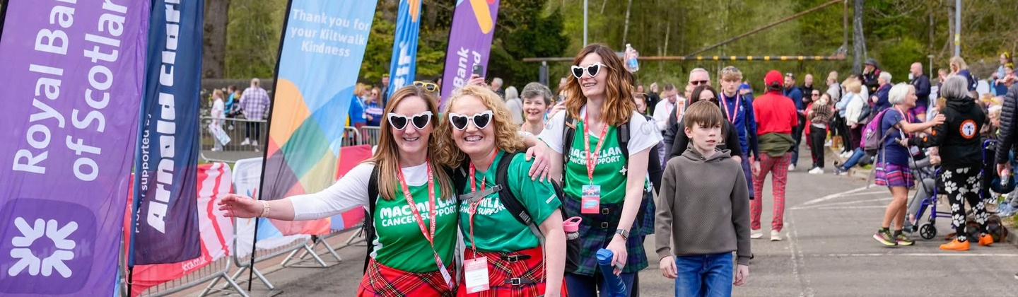 A group of people taking part in the Aberdeen Kiltwalk. Two people are walking side by side and are wearing green Macmillan branded tops and red kilts. They are walking along a paved road at the event.