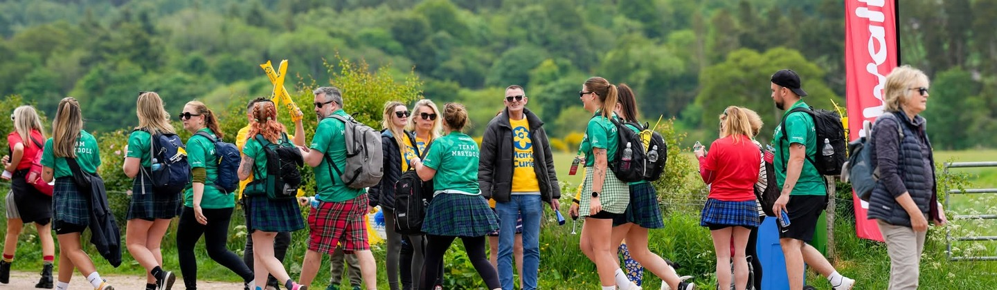 A group of people are participating in the Edinburgh Kiltwalk. They are walking along a dirt path in the countryside. Some people are wearing kilts and green Macmillan branded tops.