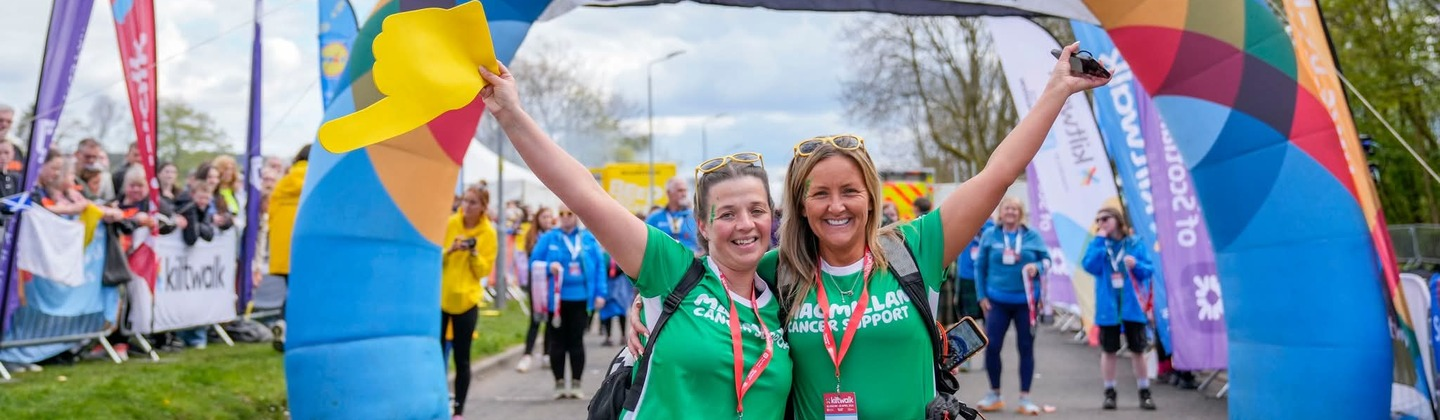 Two people are standing side by side in front of a finish balloon event arch at the Dundee Kiltwalk event. Each have opposite arms in the air and are wearing a green Macmillan top and green kilts.