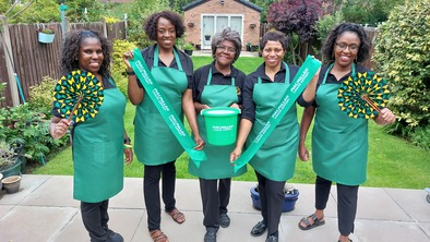Marcia is standing with a group of people in a row. They are all wearing green baking aprons. The person in the middle is holding a Macmillan donation bucket. On either side of her the people are holding a green Macmillan sash. They are outside in a garden.