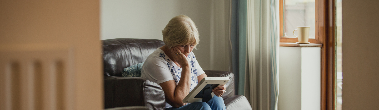An older woman is sat on a sofa and is looking down at a photo she is holding. She has one arm propped up on her leg and is holding her face. She appears sad.