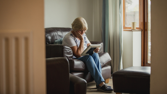 An older woman is sat on a sofa and is looking down at a photo she is holding. She has one arm propped up on her leg and is holding her face. She appears sad.