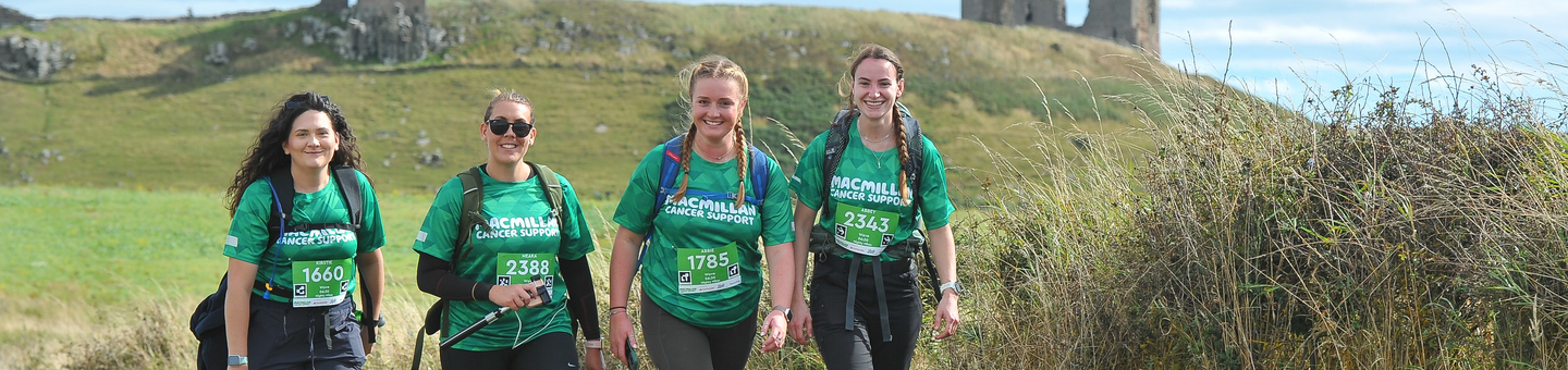 A group of four hikers are walking along a grassy path. Behind them are grassy fields and old ruins.
