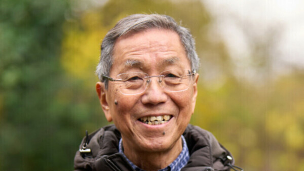 Man outdoors, smiling while wearing a dark padded jacket. The background shows greenery. 