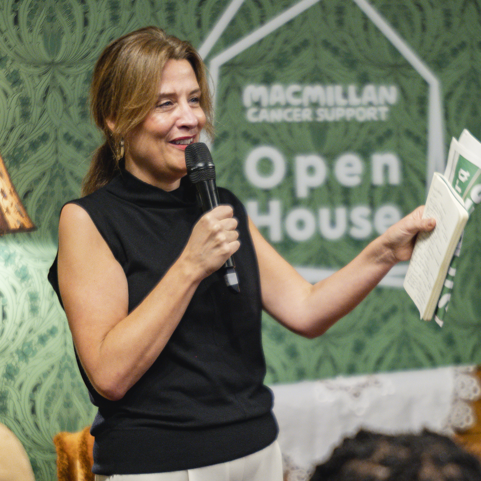Macmillan CEO, Gemma Peters, holding a microphone and reading notes at Macmillan Cancer Support Open House event.
