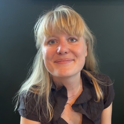 Person seated indoors against a dark green wall, wearing a short-sleeved black blouse with a ruffled neckline, with framed artwork visible above.