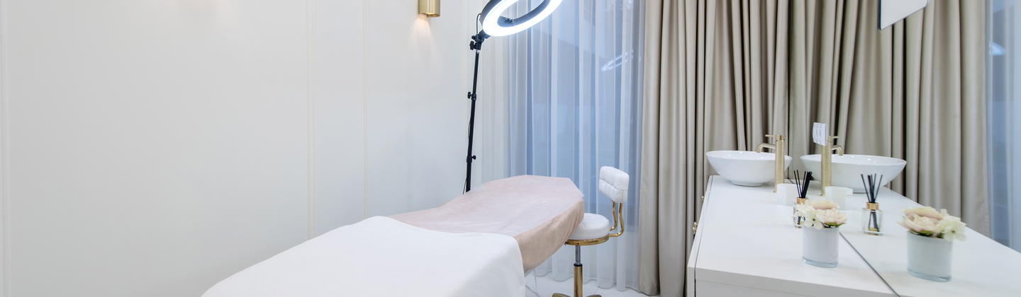 A room where beauty treatments are given. There is a table in the long table for people to lay down on in the middle. A ring light is above it. Next to the table is a counter with a sink and some decorations on it.
