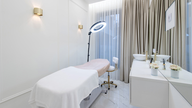 A room where beauty treatments are given. There is a table in the long table for people to lay down on in the middle. A ring light is above it. Next to the table is a counter with a sink and some decorations on it.