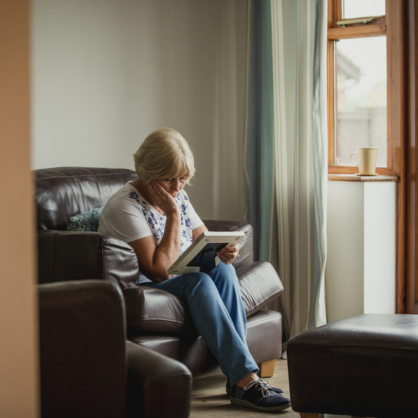 An older woman is sat on a sofa and is looking down at a photo she is holding. She has one arm propped up on her leg and is holding her face. She appears sad.