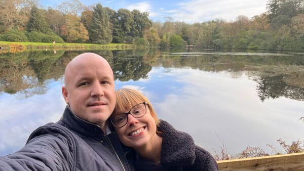 Rachel is a young woman who was diagnosed with bowel cancer. She is smiling and taking a selfie with her partner in front of a lake surrounded by trees.