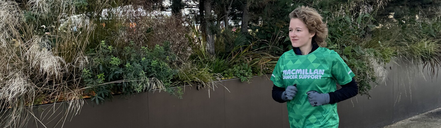 A person is running along a paved path outside. They have short curly hair and are wearing running gear including a green Macmillan branded top.