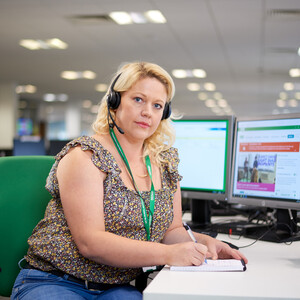 A woman working on the Macmillan Support Line. She is looking at the camera, wearing a call headset and has a notepad and pen.