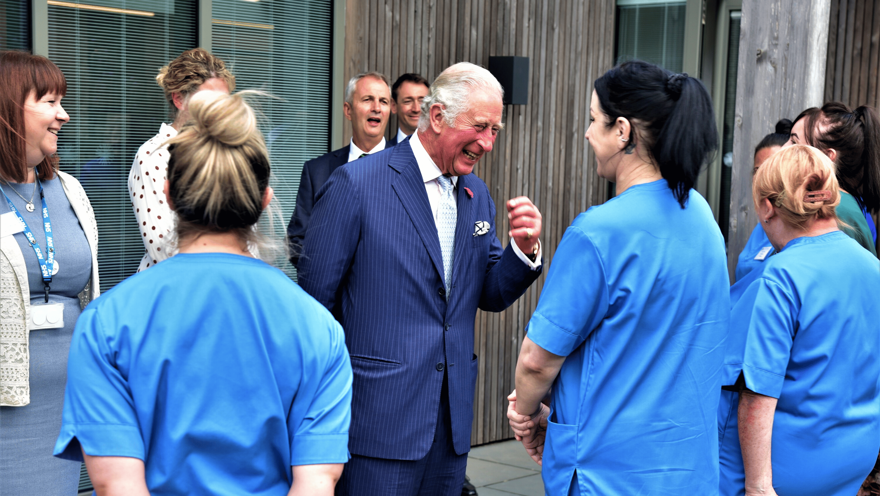 King Charles speaking with healthcare workers in blue scrubs outside a modern building.