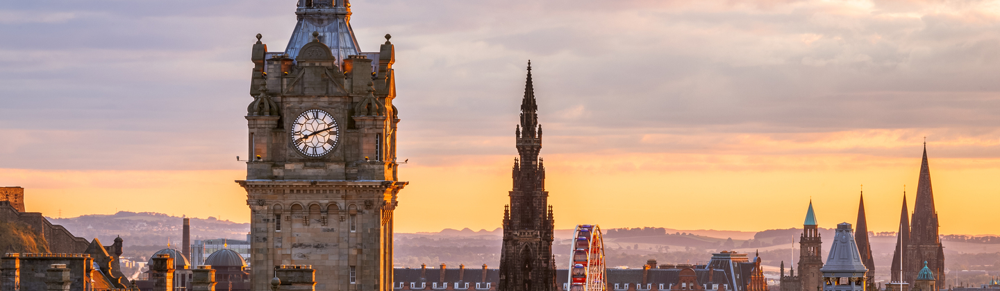 Balmoral Clocktower in Edinburgh Scotland taken from an aerial view. The sun is setting in the distance, casting an orange glow over the city. 