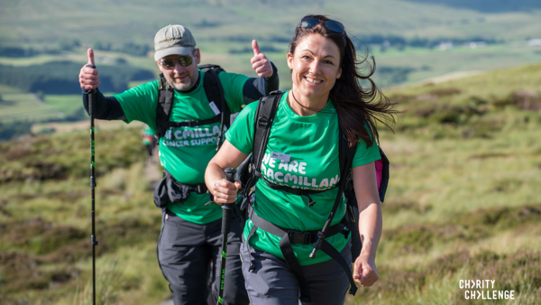Two hikers are walking along a path through a grassy field. Both of them are wearing a green Macmillan branded t-shirt and other clothes suited for hiking. One person is walking behind the other and is holding two thumbs up.