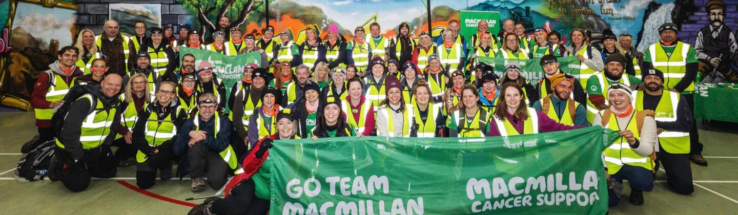 A large group of people are posing for a photo. They are wearing a mixture of hiking clothing, including head lamps. In the front, a row of people are holding up a green banner with the words 