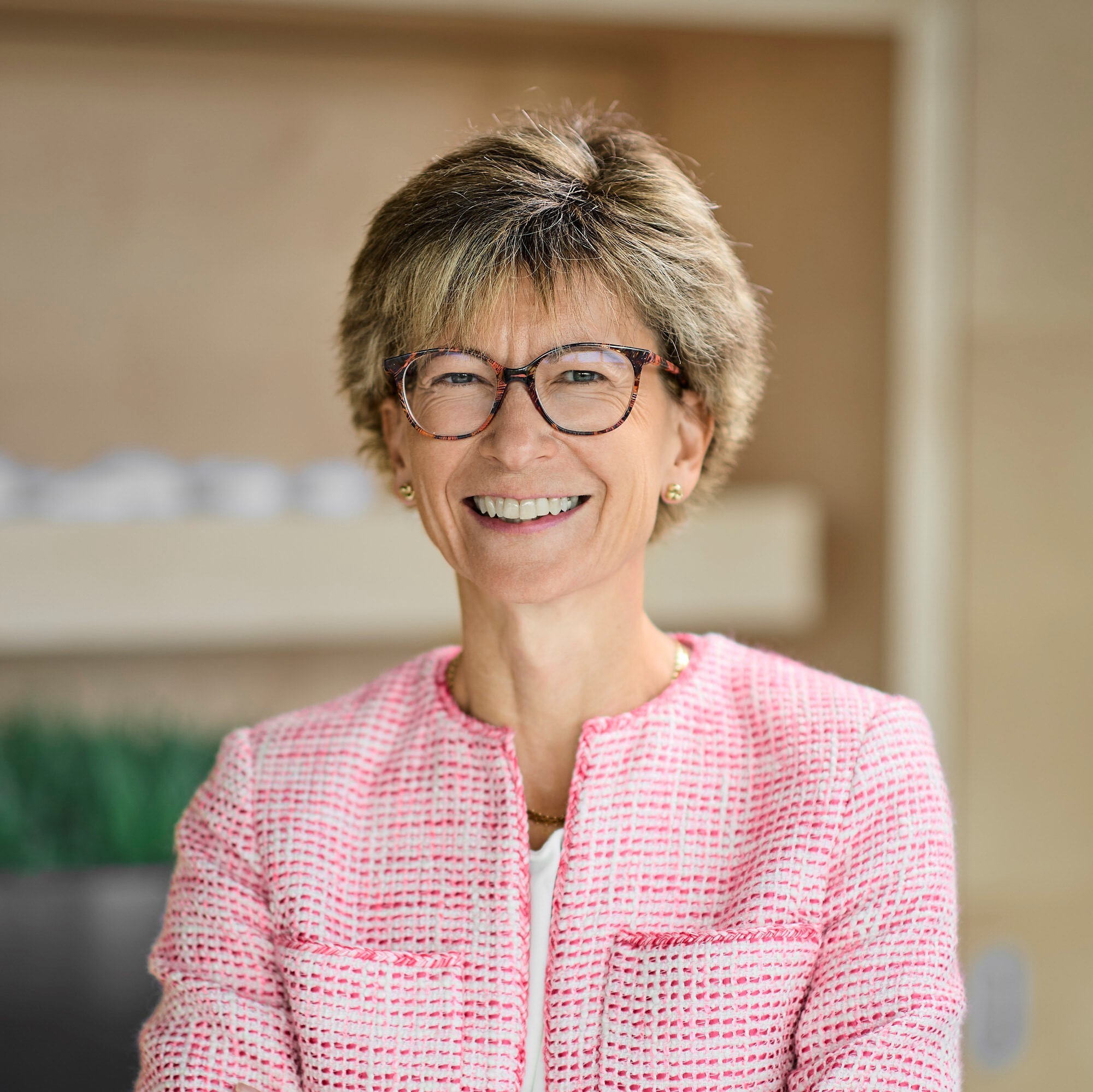Person wearing a textured pink jacket standing with arms crossed. There are shelves and plants in the background