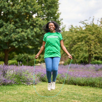 A woman is outside and is skipping a rope in a field. She is wearing a green Macmillan top, jeans, and white trainers. The rope she is skipping is green and behind her is lavender plants.