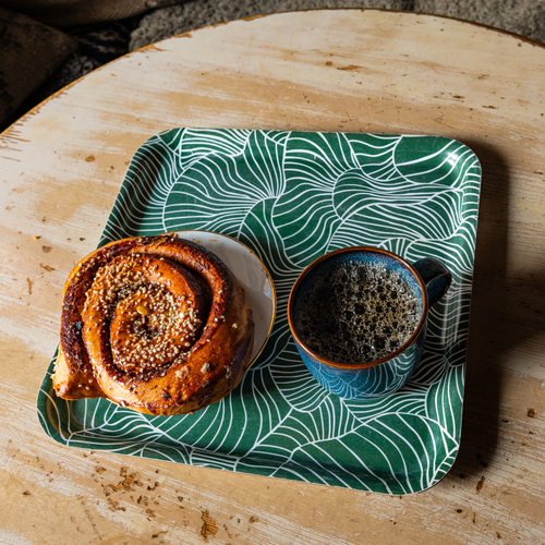 A colourful tray on a wooden table. Place on the tray is a large cinnamon bun on a plate and a dark cup of coffee.