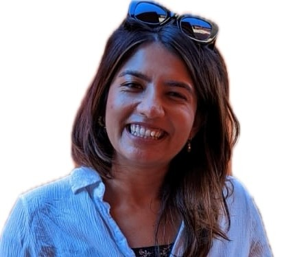 A person with long, dark, wavy hair wearing sunglasses resting on top of their head. They are dressed in a light blue, textured button-up shirt over a darker top. The background is plain white.