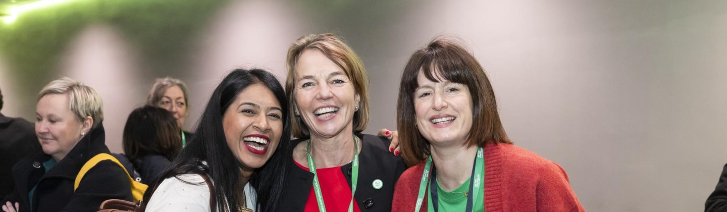 Rufsana, Claire and Fran are standing side by side. They are smiling. Rufsana is wearing a white outfit, Claire a red gown and black cardigan, and Fran is wearing a green Macmillan top, red cardigan and dark trousers.