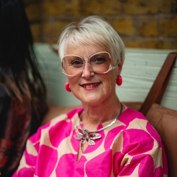 Penny has short hair and is wearing white rimmed glasses. She has on a pink and white patterned top and pink earrings. She appears to be sitting somewhere indoors.