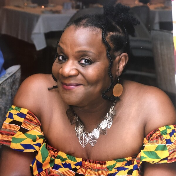 Sophia is leaning her arms on a table. She has shot dark hair that is tied up. She is wearing a colourful patterned top. Sophia has on earrings and a necklace. She appears to be indoors at an event.