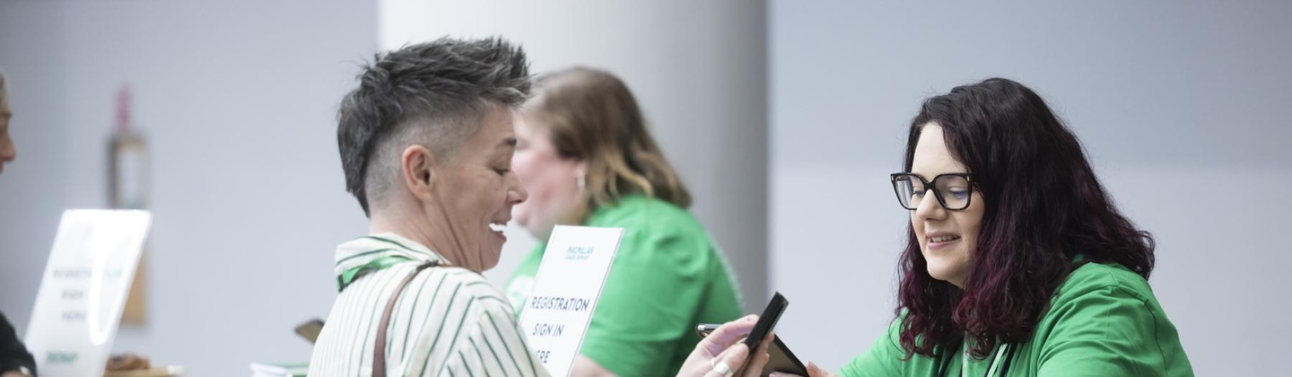 A person is standing behind a desk. They are speaking to someone who is checking their mobile. They appear to be at an event.