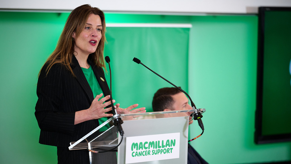 Gemma Peters, Chief Executive Officer at Macmillan, is speaking behind at a podium. She has long hair and is wearing a green top and dark jacket.