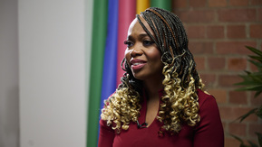 A person wearing a deep red long-sleeved top sits indoors in front of a backdrop of vertical rainbow-coloured stripes. Their hair is styled in long, dark braids with golden curls at the ends. A brick wall and green plant are visible in the background, creating a modern, vibrant setting.