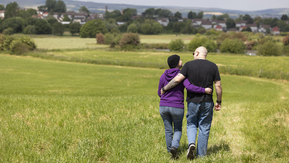 Two people walking arm in arm. They are in a field and they have their back to the camera.