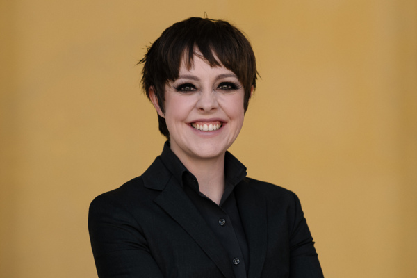 Woman with short dark hair stands in front of an orange wall. She is wearing a black suit and has her arms folded.