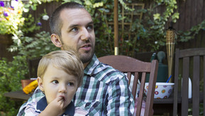 Luke is sat in the garden with his son on his lap.