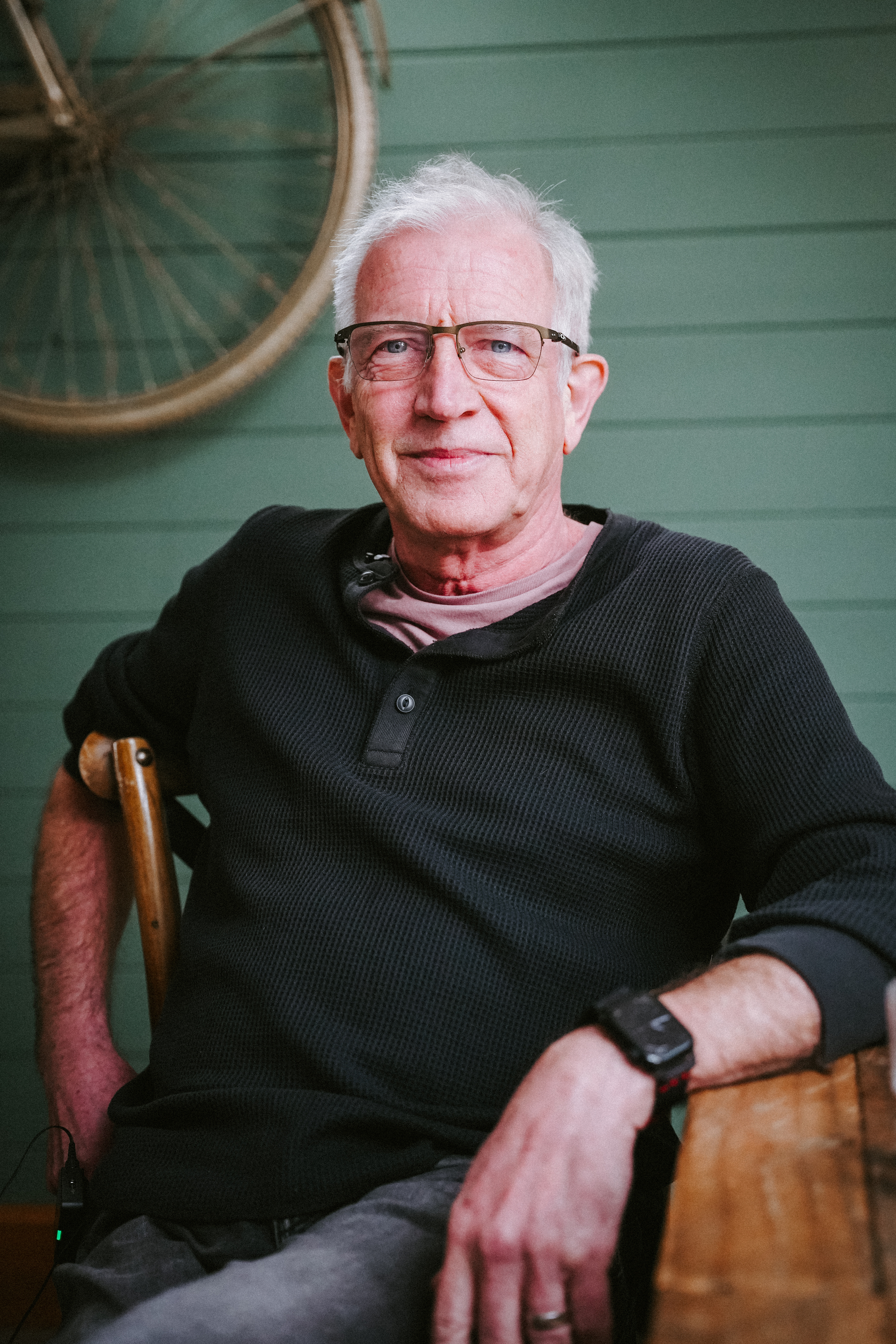 A man sitting casually at a table. A bike wheel can be seen mounted on the wall behind him