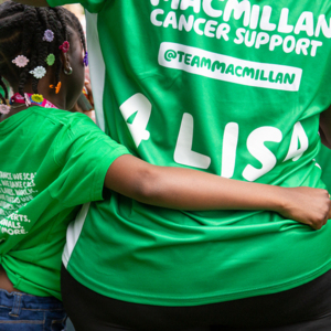 A small child with their arm around an adult's waist. The child has braids and flower hair clips in their hair. The child and the adult are wearing green Macmillan t shirts. On the back of the adult's t-shirt it says '4 Lisa'.