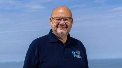 Neil is standing outside in the sunshine with the sea behind him. He is smiling and wearing glasses and a top with the North Wales Prostate Cancer Support Group name, a Welsh dragon, and a blue ribbon on it. 