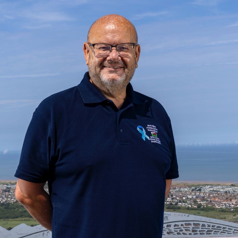 Neil is standing outside in the sunshine with the sea behind him. He is smiling and wearing glasses and a top with the North Wales Prostate Cancer Support Group name, a Welsh dragon, and a blue ribbon on it. 