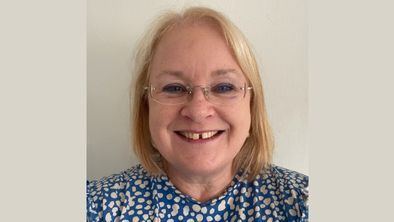Woman smiling at camera, she is wearing  a blue top with white spots. She has blonde hair and is wearing glasses.