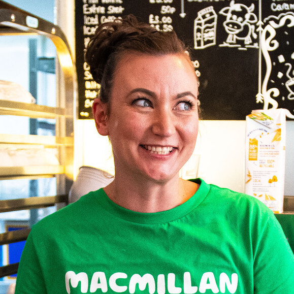 Jenny is standing and leaning against a metal table with an espresso machine on it. In front of them is a wood table with different baked items on it. There is also a Macmillan branded money bucket. The person appears to be in a café. 