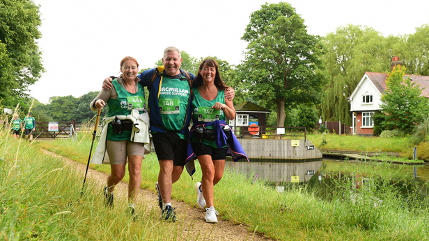 Three hikers are walking together side by side. The person in the middle has his arms around each hiker to his side. They are walking along a path in the Cotswolds.