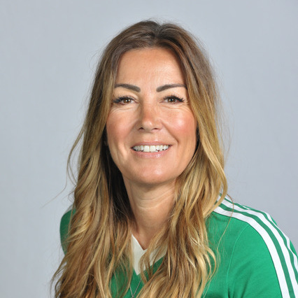 A head shot of Tania. She has long wavy brown and blonde hair. She is wearing a green top and a necklace. She is standing in front of a grey background.