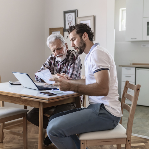 A father and son are talking to one another whilst sat at a dining table. They have a laptop open on the table and some other paperwork around them.
