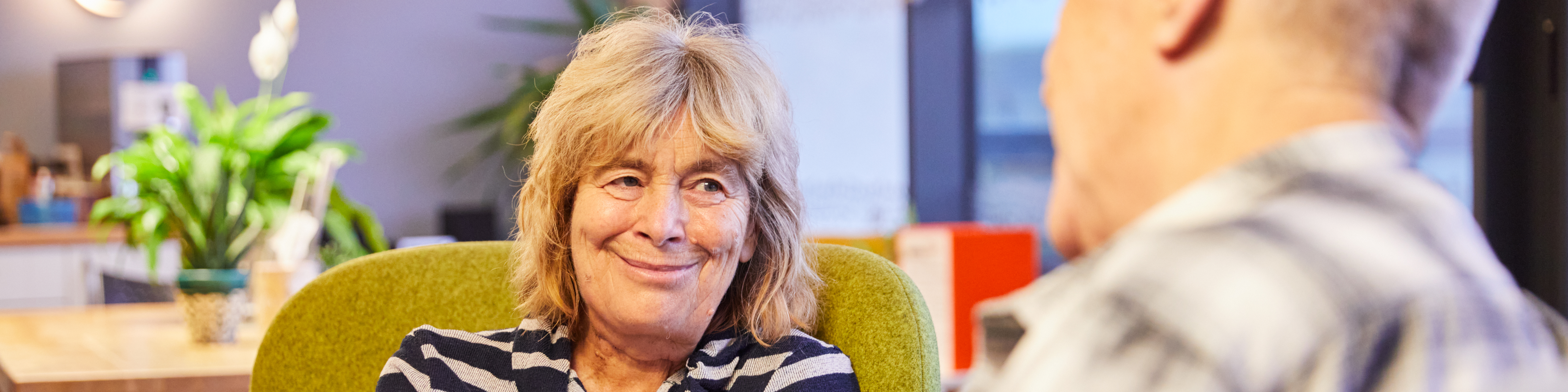 Two people sit facing each other. One person sits on a green chair, wearing a striped top, with hands resting together. The other person is seated in the foreground, slightly out of focus.