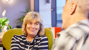 Two people sit facing each other. One person sits on a green chair, wearing a striped top, with hands resting together. The other person is seated in the foreground, slightly out of focus.