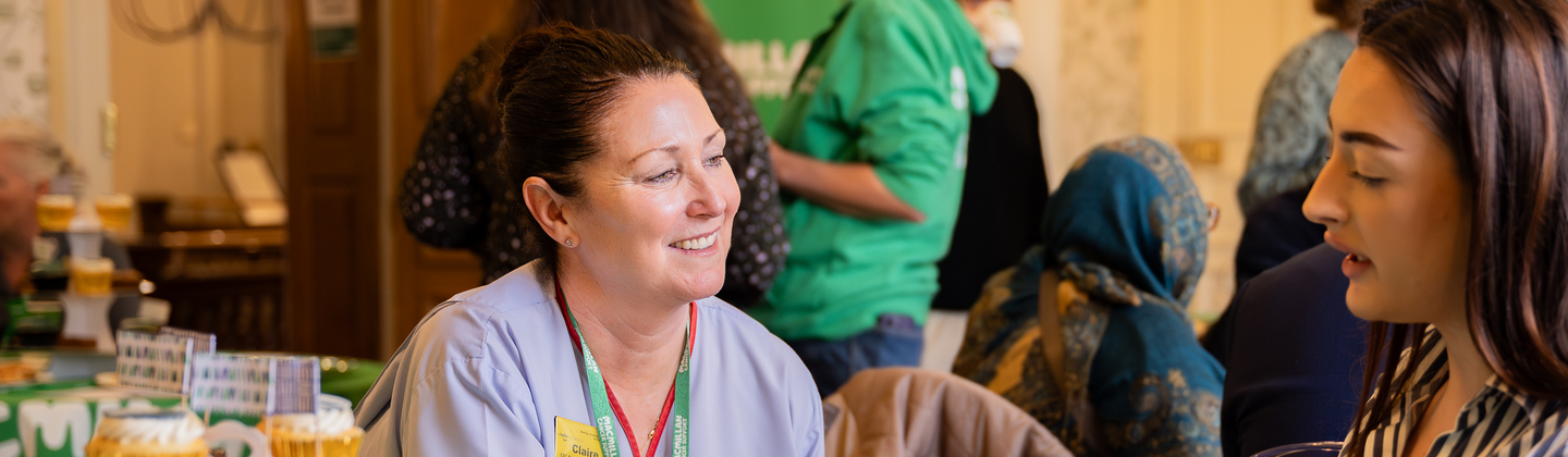 A Macmillan nurse and a Coffee Morning attendee are sat at a table and talking. On the table are different treats and drinks. They appear to be in a conference room filled with other Coffee Morning attendees.