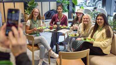 A group of people are sat around a table and attending a Coffee Morning at the Macmillan's London office. Someone is taking as selfie of the group. They all have yummy Coffee Morning treats with them.