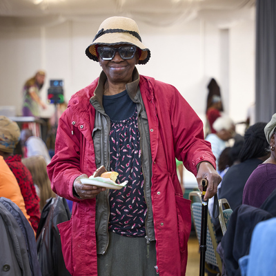 A lady is standing and smiling at the camera. The photo was taken at a Coffee Morning with the Pineapple Club in London. She has on a sun hat, sunglasses, a pin jacket, skirt and colourful top. She is holding a plate with different yummy Coffee Morning treats on it.