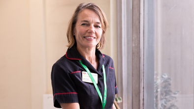 Nurse in a dark uniform with red trim stands by a large window, holding the wooden handrail. 
